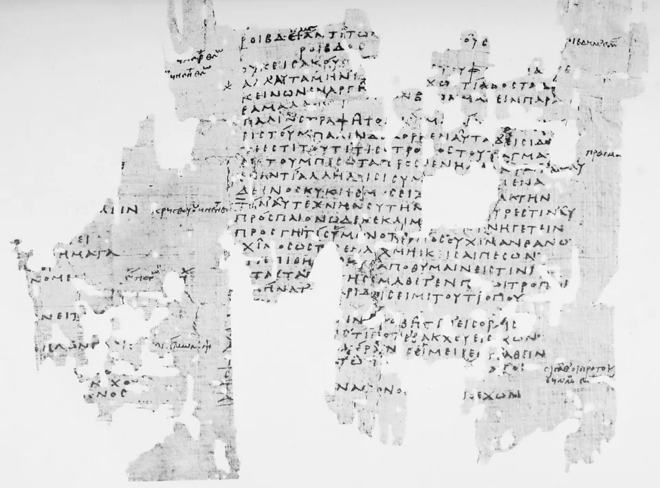 A torn sheet of papyrus with two columns of Greek capitals written by hand, many holes and missing edges, and small notes in the margins.